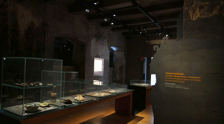 Die ehemalige Räucherkammer mit dunklen Wänden. Beleuchtete Vitrinen stehen in der Mitte des länglichen Raumes. Am rechten Bildrand befindet sich eine Metallplatte, die den Umriss einer Frau darstellt.