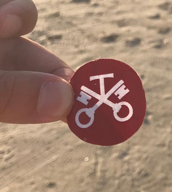 Eine Hand hält einen roten Aufklebepunkt. Im Hintergrund befindet sich ein Strand.