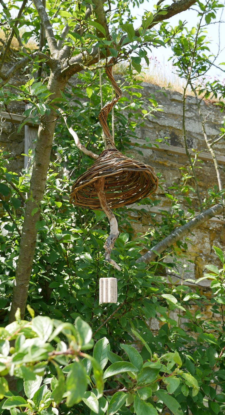 Ein Vogelcafé hängt zwischen grünen Zweigen. Es hat ein kleines rundes Dach aus Weidenruten und darunter hängt ein Stock, auf dem Vögel sitzen können sowie ein kleiner Block Vogelfutter.