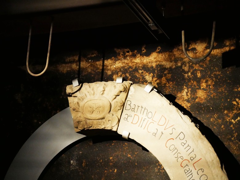 An der dunklen Wand der Räucherkammer hängen Bruchstücke des Eingangsportals des Klosters Dalheim. Von der Decke darüber ragen zwei Metallhaken in U-Form ins Bild.